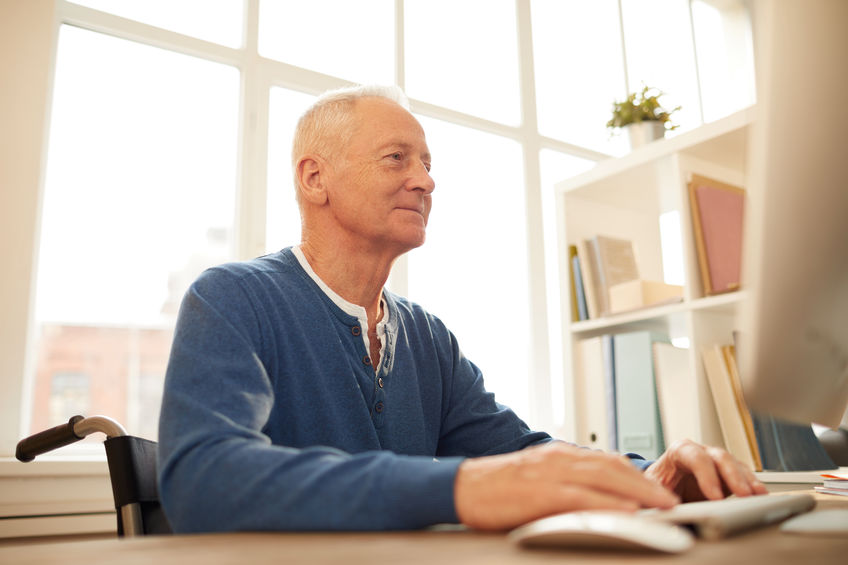 smiling older man sitting in a wheelchair using a computer at home