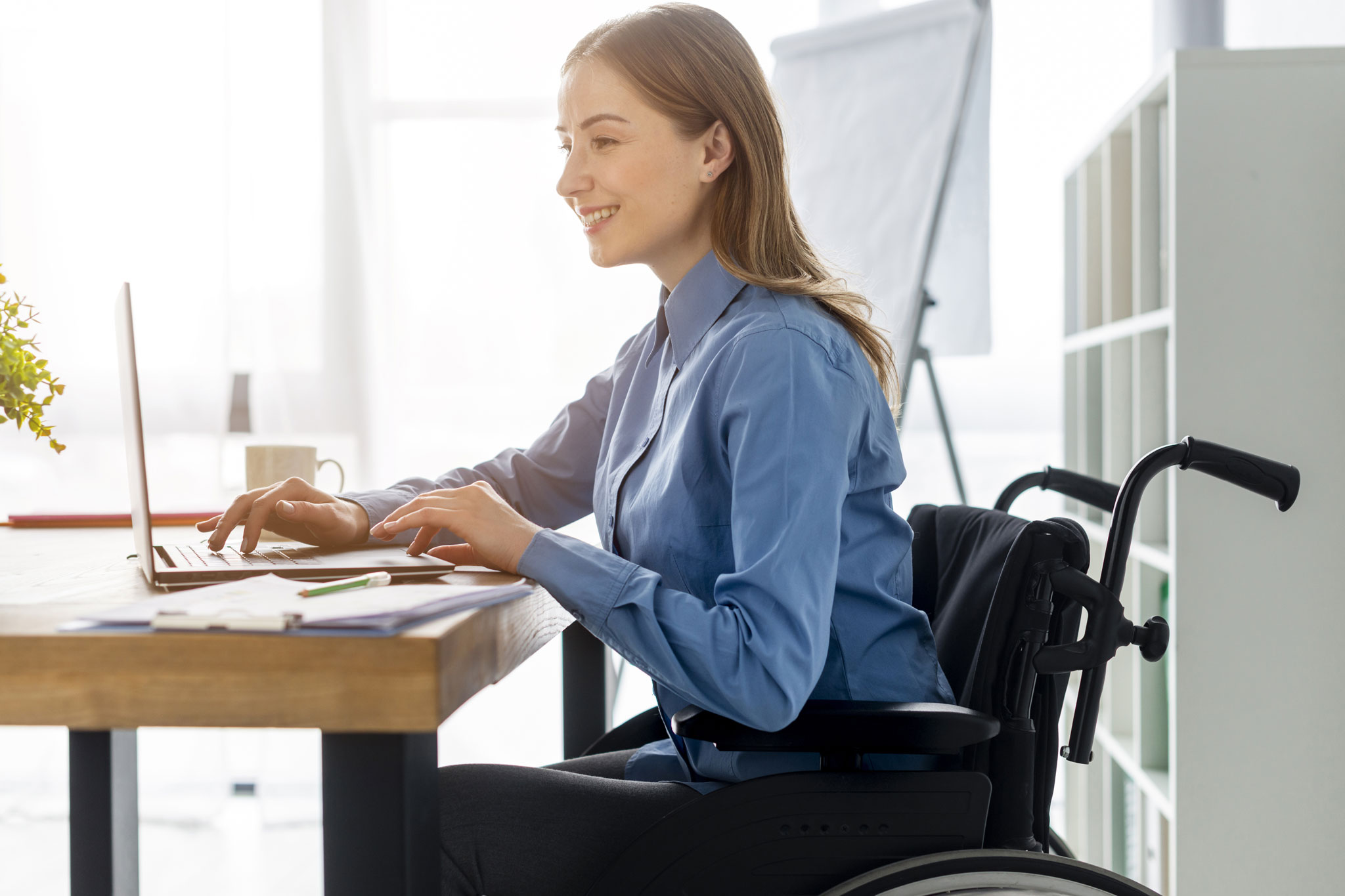 En la imagen se observa a una mujer con movilidad reducida, sentada sobre una silla de ruedas, trabajando en un portátil, aprovechando las bondades de la usabilidad y accesibilidad web.