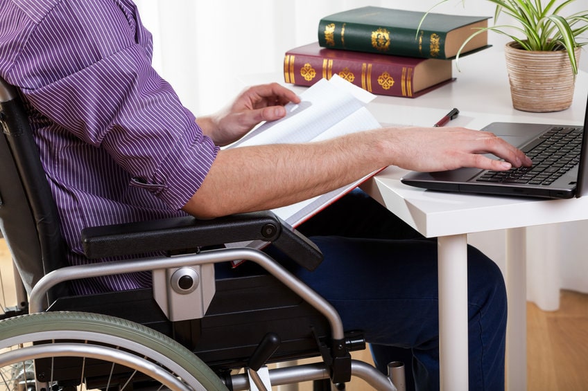Jeune homme en situation de handicap assis dans un fauteuil roulant, travaillant sur un ordinateur portable à une table.